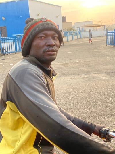 A man is depicted from a close-up angle, wearing a beanie and a tracksuit. He seems to be riding a motorcycle or scooter, holding onto the handlebars. The background shows a paved area, possibly a parking lot or street, with buildings and a fence in the distance. There is another person farther away near the center left of the shot. The image has a warm, natural light, suggesting it might be taken during sunrise or sunset.