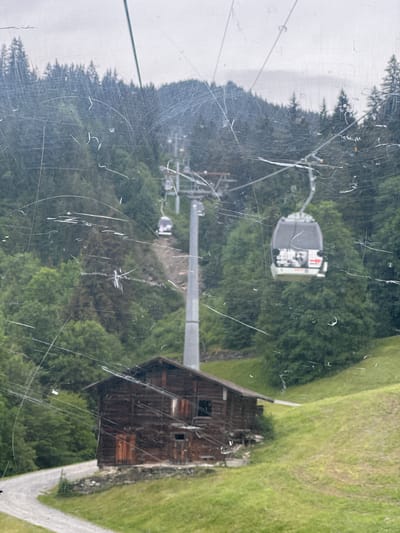 The image captures a mountain landscape viewed through a scratched window, likely from inside a moving cable car. The scene depicts a steep, tree-covered mountainside with a prominent cable car system ascending. Several cable car cabins are visible, one in the foreground featuring a graphic on its rear window. Below the cable car line, a rustic, weathered wooden cabin or barn is situated on a grassy slope, with a winding gravel road leading away from it. The environment is characterized by dense evergreen forests and a rolling, green hillside under a cloudy sky. This view is consistent with the scenery around Hasliberg Reuti, Switzerland, a known location for cable car transport in a mountainous region.