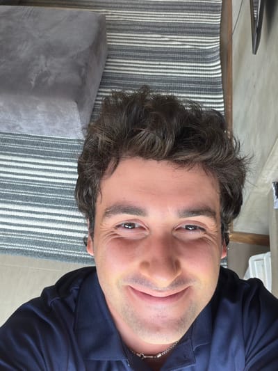 A man poses close to the camera. He has dark brown hair with light skin. He is wearing a dark blue collared shirt. A silver chain necklace is visible around his neck. Behind him is a gray and white striped rug and a gray cushioned object. The image is rotated sideways.