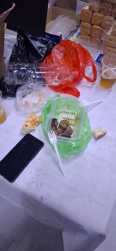 A messy, tabletop scene, likely in Arys, Kazakhstan, depicts the aftermath of a meal. A white tablecloth, marked with blue crayon scribbles, covers the surface. In the center, a green plastic container holds several pieces of grilled meat, alongside a white plastic fork. To the left, a piece of flatbread is visible, and further left, a black smartphone with a cracked screen lies on the cloth. A clear plastic bag contains remnants of food, and a large red plastic bag lies open. In the background, empty plastic bottles and other discarded packaging are scattered. To the right, a glass of amber liquid, likely beer, sits near a stack of packaged cookies. The overall impression is one of disarray and consumption.