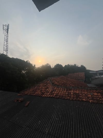 A view from a rooftop in Pasir Gadung, Indonesia, looks towards a hazy sunrise. The foreground is dominated by two distinct rooftops: one corrugated, dark gray roof in the lower section, and another roof made of reddish-brown tiles above it. The tiled roof shows some damage, with a few tiles dislodged. Beyond the rooftops, a dense line of dark green trees forms a canopy. To the left of the trees, a tall, slender cell tower stands against the pale sky. The sun appears as a small, bright orb low in the sky, partially obscured by clouds and haze, casting a diffused light. In the background, a few buildings are visible through gaps in the trees. The overall impression is of an urban or semi-urban environment at dawn.