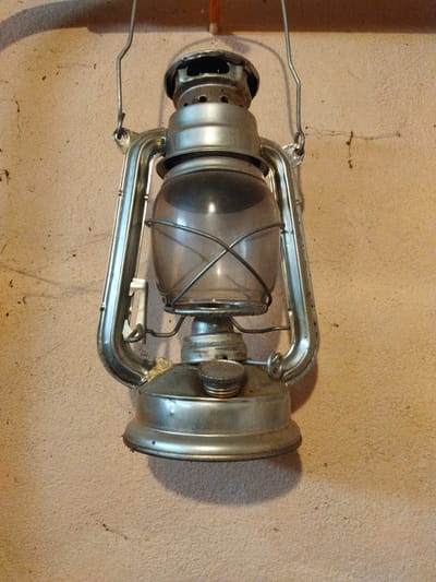 A vintage kerosene lantern hangs on a textured, beige wall. The lantern is made of metal and glass, with a handle at the top for carrying or hanging. It appears to be old and slightly tarnished. The wall has a rough, stucco-like finish with some small dark spots, possibly dirt or imperfections. The lighting suggests an indoor setting, possibly a rural dwelling or an older building. The image was taken in Klishino, Russia. There are no people present.