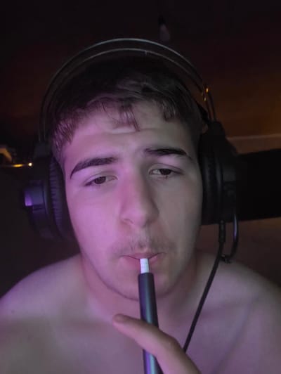 The image is a close-up, selfie-style shot of a young Caucasian male, appearing to be in his late teens or early twenties. He is wearing large, black over-ear headphones and is holding a dark cylindrical electronic device with a white tip to his mouth, suggesting he is using it. The lighting is dim, with purple and pink hues illuminating his skin, particularly on his face and upper chest, indicating an artificial light source, possibly from a monitor or LED lighting. His expression is neutral. He is shirtless. The background is dark and indistinct, with hints of warm light in the upper right corner, suggesting an indoor environment, possibly a room. There is no indication of the city of Aleksandrovo, Bulgaria, or any urban environment within the image.