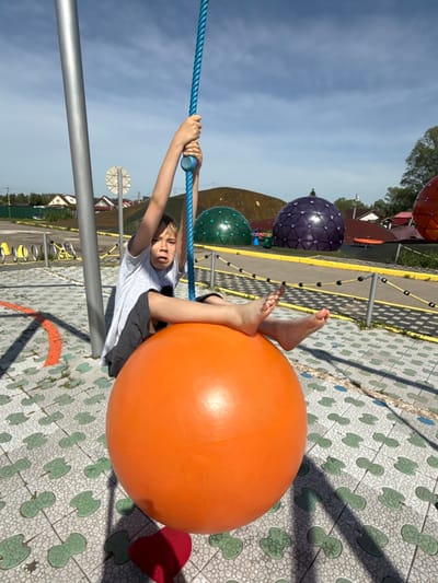 A child is on playground equipment in Синьково, Russia. The child is suspended, lying with his bare feet on an orange ball and his hands gripping a blue rope supported by a metal pole. The playground surface is a patterned tile. Behind him are additional spherical playground elements in purple, green, and orange. In the background, there is a cluster of houses and trees under a partly cloudy sky.