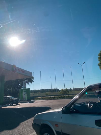 The image depicts a sunlit day at a gas station identified as "amic ENERGY" in Herasymivka, Ukraine. The foreground features the hood and side of a silver car, with its steering wheel and side mirror visible through the windshield. In the midground, a black SUV is parked at a green gas pump. The gas station canopy, also green and white, is visible with the "amic" logo prominently displayed. Tall, thin poles extend upwards, and streetlights are visible in the distance. The background consists of a treeline and a field under a clear blue sky, with the sun shining brightly and creating lens flare. The scene appears to be captured from inside a vehicle, looking out.