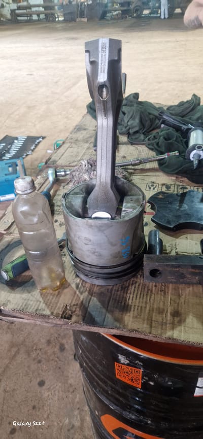 The image depicts a disassembled engine piston and connecting rod resting on a wooden surface. A plastic bottle with a yellow liquid is positioned to the left of the piston. Various tools, including a wrench set and what appears to be a pneumatic tool, are scattered around the central components. In the background, a workshop environment is visible with machinery and possibly other vehicles. The overall scene suggests mechanical work or repair in progress. The text "Galaxy S22+" is visible in the bottom left corner of the image, indicating the device used for capture.