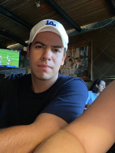 This image shows a young adult male positioned in the foreground of a public establishment in Boca del Río, Mexico. He wears a white baseball cap with a blue "LA" logo and a dark blue t-shirt, looking directly at the camera with a neutral expression. His forearms are visible across the lower portion of the frame. In the background, to the left, a television screen displays an American football game. To the right, a framed painting depicting a social gathering with multiple figures seated around a table is mounted on a dark wall with a diagonal grid pattern. Partially obscured by the foreground subject's right arm, another individual with dark, wavy hair and a light-colored shirt is visible, seated and looking away from the camera. The ceiling is composed of dark wooden slats or beams.