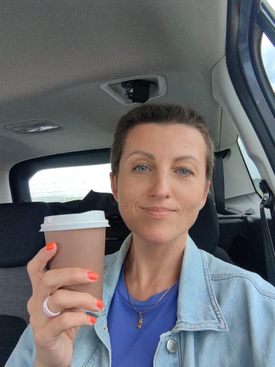 A woman with short brown hair and blue eyes is sitting in the passenger seat of a car, holding a brown paper coffee cup with a white lid in her right hand. She is wearing a blue t-shirt and a denim jacket. Her fingernails are painted bright orange, and she is wearing a light pink ring on her left hand. The interior of the car is visible, including the grey headliner and a window showing a slightly overcast sky and a distant landscape. The image appears to be a selfie taken by the woman. The location is identified as Хаценчыцкі сельскі Савет, Belarus.