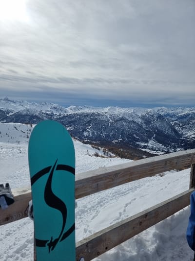 The image depicts an alpine winter landscape captured from a high vantage point in Cesana Torinese, Italy. In the left-center foreground, a turquoise snowboard with a black, stylized 'S' or serpentine emblem is positioned vertically. A weathered wooden railing, composed of two horizontal planks, extends across the lower mid-ground. On the left portion of this railing, a gray and black patterned glove is visible. A segment of a blue garment is partially visible on the far right, positioned behind the railing. The immediate ground surface is covered in textured snow. The background features extensive snow-capped mountain ranges under an overcast sky, with a distinct bright light source in the upper left quadrant. The distant mountain slopes display patterns of snow, dark coniferous forests, and areas of sparse deciduous trees. No individuals are explicitly depicted, but the presence of the snowboard, glove, and garment indicates recent human activity.