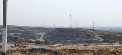 The image shows an open-pit mine in Margilan City, Uzbekistan, under a hazy sky. The foreground is dominated by dry, yellowed grass and rough terrain, with a concrete utility pole on the left side. The middle ground features a large excavation with layered rock formations and piles of gravel or sand, suggesting ongoing mining operations. Several dirt tracks wind through the mine. In the background, numerous high-voltage electricity pylons march across the landscape, indicating significant power infrastructure. The distant horizon is obscured by haze, with faint outlines of buildings and trees suggesting a populated area beyond the mine. No people are visible in the image.
