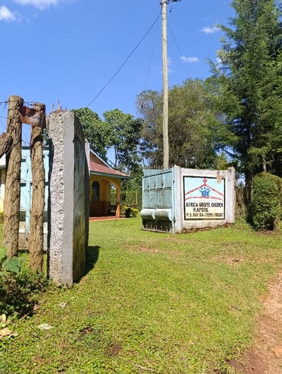This outdoor image captures the entrance to the Africa Gospel Church Kapsiya, set against a clear blue sky. The scene is static, with no people present or discernible activity.
In the foreground, a neatly trimmed, vibrant green lawn dominates the ground, with a reddish-brown dirt path visible on the right. The entrance features a concrete gate post on the left, behind which a wooden fence post with barbed wire and a small, partially legible, faded sign are visible. To the right, a light-blue metal gate is partially open, revealing a glimpse of the church property beyond.
A prominent, rectangular sign for the "Africa Gospel Church" is affixed to a concrete block on the right side of the entrance. The sign displays a logo with a stylized cross and human figures, and the text below it clearly reads: "AFRICA GOSPEL CHURCH", "KAPSIYA", and "P.O. BOX 84-20205 SOSIOT".
Beyond the entrance, a section of the church building is visible, characterized by a light-colored wall, arched window frames, and a reddish-brown roof. A tall, light-colored wooden utility pole stands centrally in the midground, extending high into the sky with several electrical wires attached. Lush green trees of various types form a dense background, suggesting a verdant environment.
The bright sunlight indicates it is daytime, casting clear shadows on the ground and elements. The sky is a vivid blue with a few sparse white clouds. The visible text on the church sign explicitly names "Kapsiya," aligning with the given location context of Kaplelwo, Kenya.
