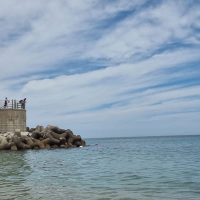 This is an image of the coastline in Gangneung-si, South Korea. In the foreground, the clear, blue ocean laps gently against the shore. In the midground, a concrete structure with railings stands on a breakwater, adorned with relief carvings. Two people are visible on top of this structure, one facing away from the camera, and another leaning over the railing, looking down. A pile of tetrapods, large concrete structures designed to protect coastlines from erosion, sits to the right of the concrete structure, partially submerged in the water. Further out in the ocean, a small red buoy is visible. The sky above is a mix of blue and white clouds, with a light breeze creating wispy patterns. The overall scene depicts a peaceful day at the beach, with a focus on the man-made elements of coastal defense and a natural, serene environment.