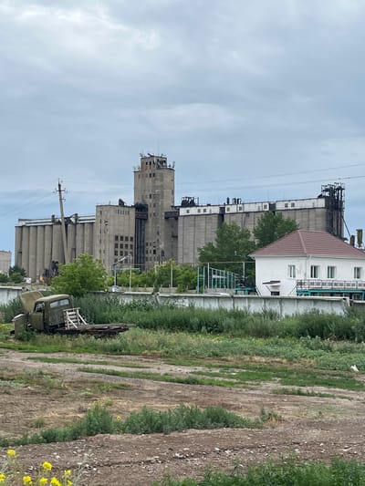 The image shows a weathered industrial facility, likely a grain elevator, under a cloudy sky in Chelgashi, Kazakhstan. The main structure is a tall, concrete building with multiple silos and a central tower, showing signs of age and neglect with visible staining and some missing elements on the roof. To the right of the industrial complex, a small, white, two-story house with a reddish-brown tiled roof stands out, appearing more maintained. In the foreground, an old, green, military-style truck sits in a field of overgrown grass and weeds. The truck is damaged, with its hood up and the bed empty except for some wooden planks. The surrounding area is a mix of dry, dusty ground, patches of green vegetation, and scattered debris, suggesting an abandoned or disused industrial zone. The overall atmosphere is one of decay and abandonment, juxtaposed with the presence of a functioning residential building.