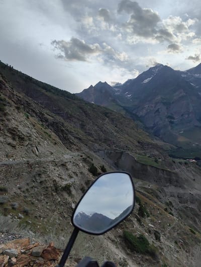The image depicts a mountainous landscape in the vicinity of Gushal, India, taken from the perspective of a motorcycle rider. In the foreground, a motorcycle's side mirror reflects a portion of the scene, showing snow-capped mountains under a cloudy sky. The main view consists of steep, rocky mountain slopes with sparse vegetation. A narrow, winding road is visible carved into the mountainside. Further in the background, more rugged peaks rise, some with patches of snow. The overall environment suggests a remote, high-altitude region.