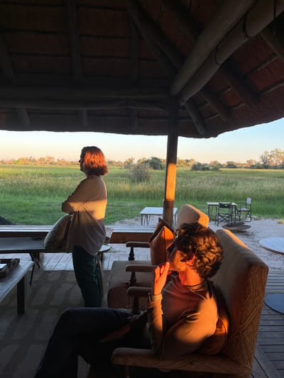 The image, taken in Khwai, Botswana, shows an outdoor scene with three people situated on a wooden porch structure under a thatched roof. The background features a flat landscape with green vegetation under a clear sky.
A man is seen on the right, sitting in a patterned armchair and looking downwards at something in his hands. To his left, a woman stands near the edge of the porch, her gaze directed towards the landscape. Another person, only partially visible with dark clothing, is on the right side of the frame. Outdoor furniture is visible in the background, including tables and chairs. The scene is illuminated with a warm, late-day light.