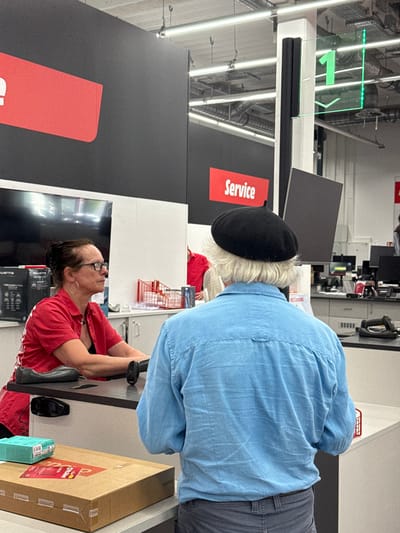 An indoor shot shows a store's service area. A woman wearing glasses and a red shirt sits at the counter scanning items. A customer in a blue shirt and beret waits behind her. A "Service" sign is visible. Overhead, a green arrow is illuminated above a row of lights. The environment appears to be a retail store, with a check out area. A product box is on the right corner of the counter.