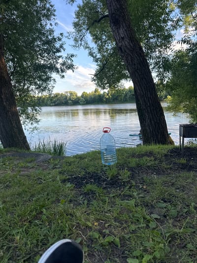 The image shows a view of the Klyazma River in Il'inskoye, Russia. In the foreground, there is a large plastic bottle of water with a red cap, sitting on the ground next to a fire pit that has been used. The ground is covered in grass and dirt. Two large trees are on either side of the frame, with their branches extending upwards and creating a canopy of leaves. The river is calm with ripples on its surface, and there are some plants growing in the water. In the background, a line of trees and greenery forms the opposite bank of the river. A person is visible in the middle ground, standing on a paddleboard in the river. A barbecue grill stands to the right of the frame, near the riverbank. The sky is partly cloudy with a mix of blue and white. The overall scene depicts a relaxed outdoor setting by the river.