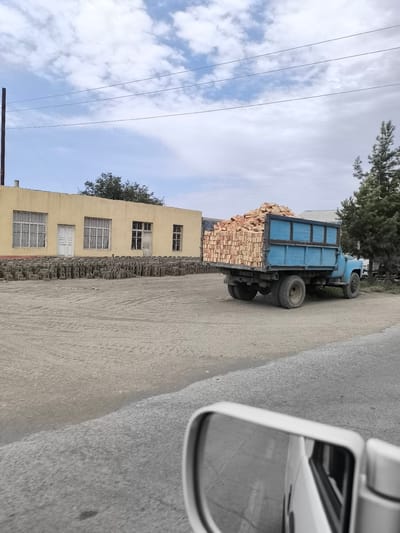 In the city of Kichik-Turk, Uzbekistan, a blue truck is parked on a dirt road, its bed loaded with bricks. In the background, a long, yellow building with several windows and doors stands. Piles of bricks are stacked neatly in front of the building. The sky is a mix of blue and white clouds, and a few trees are visible. The overall scene suggests a construction or manufacturing site.