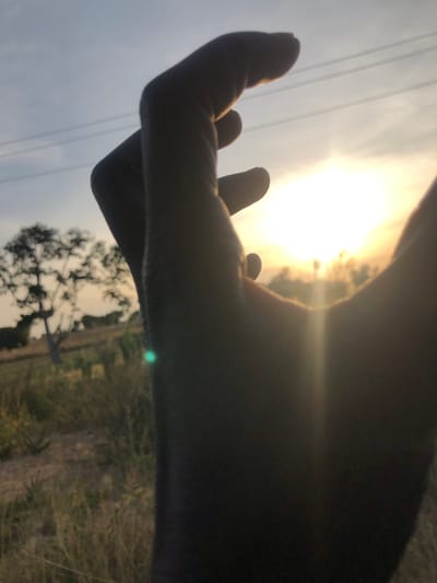 The image depicts a human hand, silhouetted in the foreground, reaching upwards towards a bright sun positioned centrally in the upper right quadrant of the frame. The hand, likely male given its size and robust appearance, is partially obscuring the sun, causing lens flare visible as a bright green dot near the bottom left and a vertical light streak. In the mid-ground, a lone, deciduous tree with sparse foliage stands on a slightly elevated, dry, grassy plain. Faint lines resembling power lines are visible against the sky in the upper left. The sky exhibits a gradient from light blue at the top to bright yellow and orange around the sun. The environment is consistent with a rural or semi-rural landscape in Barakin Kuru III, Nigeria, at either sunrise or sunset.