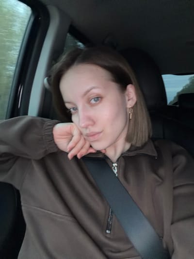 A woman is in a car, wearing a seatbelt and a brown zip-up jacket. Her hand is near her face. Outside the car window, it appears to be raining or wet. The image does not provide any information about the location of Слобода, Russia.