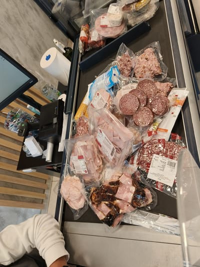 This overhead shot captures a bustling supermarket checkout or deli counter scene. The primary focus is a collection of various vacuum-sealed deli meats and charcuterie laid out on a dark conveyor belt. These include numerous packages of sliced ham, salami (some labeled "Milano"), diced bacon, and other processed meat products. Many packages feature price labels, barcodes, and product information, including "TESCO" on one item and "ská slanina" (Czech for "smoked bacon") on another, indicating a Czech retail environment. Visible price points include "15,0 Kč" (Czech Koruna) for a "0.175 kg" item, and an expiration date of "01.07.2026" on another.

To the left of the conveyor belt, various checkout accessories are visible. These include a computer monitor displaying the word "livama," a black receipt printer, a barcode scanner, and a roll of white paper towels with a blue cap. A clear plastic water bottle, partially visible, shows the brand "Dona." A person's arm, wearing a white long-sleeved top, is visible in the bottom-left corner of the frame, suggesting they are either assisting with the transaction or waiting for their turn. The background features a light grey textured wall and wooden slatted panels. The image depicts the process of scanning or preparing a customer's purchase of deli items at a grocery store, consistent with the Jesenice, Czechia location context.