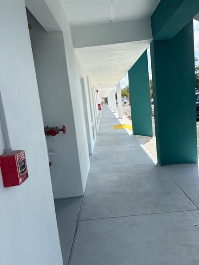 This is a high-angle, outdoor, daytime photograph taken in South Bradenton, USA. The scene depicts an architectural structure with multiple levels, characterized by white concrete walls and teal-colored beams forming a canopy overhead. The perspective is looking upwards and along a narrow passageway.

In the foreground, a red fire alarm pull station is visible on a white ledge, with the word "FIRE" prominently displayed. Further along the wall, a red fire hydrant with a visible chain is mounted. The walls are clean and appear to have a fresh coat of white paint. The ground appears to be a concrete walkway.

The background shows a glimpse of the sky with scattered clouds, suggesting fair weather. Trees and some distant structures are visible, hinting at an urban or suburban environment. The overall impression is one of an industrial or institutional building, possibly a parking garage or a service area, designed with functional elements like fire safety equipment. There is no discernible activity or people in the image, and the scene conveys a sense of stillness. The lighting suggests it is daytime, likely morning or afternoon, with natural light illuminating the scene.