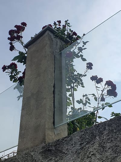 This is a close-up, low-angle shot of a concrete pillar, partially obscured by a clear glass panel. Dark red roses with green leaves are growing around and behind the pillar, some reaching towards the sky. The sky is a pale blue with some clouds. A rough, textured concrete wall is visible in the foreground. The scene is outdoors, and no people are present. The location in the city of Mukhatskaro, Georgia is not identifiable from the image.