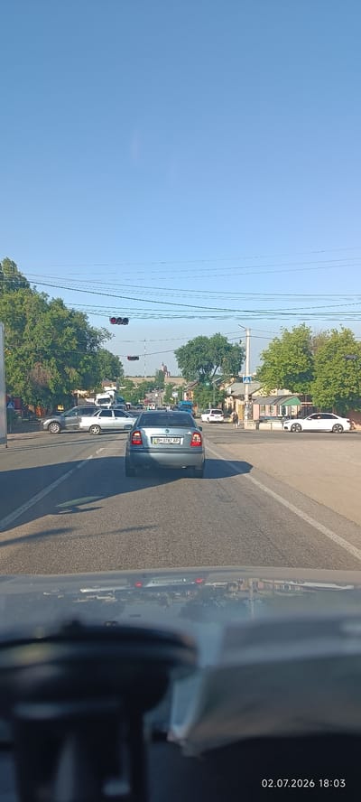 This is a vertical, eye-level shot taken from inside a vehicle, looking forward towards a moderately busy intersection. The sky is a clear, bright blue, indicating a sunny day. The time stamp in the bottom right corner reads "02.07.2026 18:03", suggesting it is early evening in the summer.
In the foreground, the dashboard and windshield of the vehicle are visible, creating a somewhat distorted reflection of the road ahead and surrounding buildings. The main subjects are the vehicles on the road. A light blue sedan with license plate "BH 0787 AP" is in the center of the frame, directly in front of the camera. To its left, a silver sedan and a gray SUV are stopped at the intersection. To the right, a white coupe is in the process of turning.
The scene is a typical urban or suburban intersection with paved roads and sidewalks. Buildings line the sides of the road, some appearing to be residential homes with pitched roofs and windows, others commercial structures. A tall traffic light with three red lights illuminated hangs over the intersection, indicating that traffic is stopped. Power lines crisscross the sky. Mature green trees with dense foliage are visible on both sides of the road, casting long shadows on the asphalt. In the distance, the landscape appears slightly hilly with more trees and buildings.
The overall mood is one of everyday activity, with traffic waiting at a light. There are no people prominently visible in the cropped sections, but the presence of multiple vehicles implies a normal flow of traffic. The setting is consistent with a street scene in a town or city, possibly in Ukraine given the location context provided. The lighting suggests a late afternoon or early evening sun, creating a warm glow on the scene.