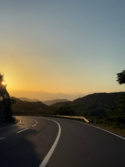 The image depicts a scenic view of a winding mountain road at sunset. The road, paved with asphalt, curves from the left foreground towards the right middle ground, marked by white dashed lines and a solid white line on its outer edge. A yellow guardrail runs along the right side of the road. The setting sun casts a warm, golden glow on the horizon, illuminating a series of layered mountain ranges that recede into the distance. The sky transitions from a vibrant yellow near the sun to a clear, pale blue at the top. On the left side of the road, a rocky cliff face is visible, partially obscured by foliage and the bright sun. Lush green trees and vegetation cover the slopes of the mountains. The overall atmosphere is serene and natural. The location is identified as Gornja Brova, Montenegro.