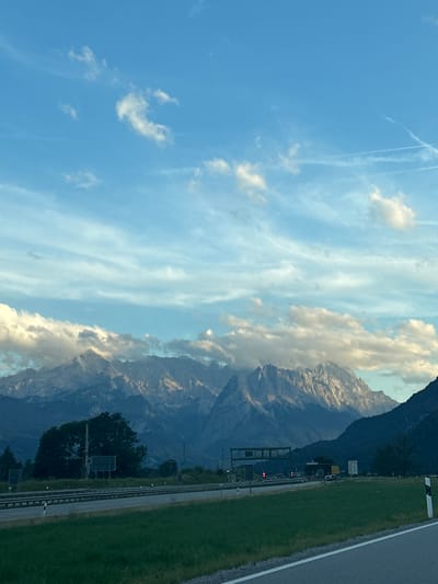 The image shows a scenic view of a highway with mountains in the background. The highway is divided into multiple lanes, with a guardrail separating the lanes. The road is bordered by a grassy median. In the distance, a range of mountains is visible, with some of them covered in clouds. The sky is blue with some scattered clouds and contrails from an airplane. The lighting suggests it is either early morning or late afternoon. There are no people visible in the image. There are some vehicles on the highway and signs. The scene appears to be in a rural or semi-rural area. This location is likely near Farchant, Germany, given the characteristic Alpine landscape.