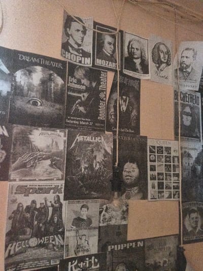 A section of an indoor wall in the city of Dogsenang Gorphoog, Bhutan, is densely covered with an informal collage of approximately 20-30 monochromatic paper printouts, posters, and flyers. These items are attached using thin, light-colored strings running horizontally and vertically across the display. The content predominantly features musical themes. Visible subjects include portraits of classical composers such as Frédéric Chopin, Wolfgang Amadeus Mozart, Johann Sebastian Bach, Antonio Vivaldi, and Pyotr Ilyich Tchaikovsky, displayed on smaller, rectangular prints in the upper section. Below these are various rock and metal band posters and flyers, including a "DREAM THEATER" poster featuring a large eye in a natural setting; an "Eric Johnson" concert flyer for "Boulder Theater" dated "Saturday, March 27"; a "METALLICA" poster depicting a roped statue; a "SEPULTURA" poster with a stylized, tribal-like face; a "QUEEN" poster displaying a grid of numerous small images; a "HELLOWEEN PUMPKINS UNITED" poster featuring a band lineup, under a "ROCK MAGAZINE Spark" title; and posters for "Cruelty And The Beast" (Cradle of Filth) and "BUCKETHEAD," along with less distinct references to "PUPPEN" and a raised fist symbol. The printouts vary in clarity, some appearing faded or grainy, and are arranged in an overlapping, haphazard manner against the plain, light brown wall.