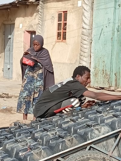 A daylight street scene in Gayawa, Nigeria, depicts two individuals in the foreground and mid-ground. An adult male, wearing a black t-shirt and dark trousers with a patterned stripe on the left leg, crouches over numerous black, rectangular plastic containers with screw-on caps, which are tightly packed on a metal-framed transport structure. He extends his right arm across the containers, while his left hand is also near them, and he looks towards the right. To his left and slightly behind, a young female, clad in a dark grey hijab and a long dark dress over a patterned garment, stands barefoot. She looks down at a red rectangular object held open in her hands. The background consists of a light-colored building featuring a grey wooden door on the left and a small reddish-orange framed window. To the right, a segment of a light green door/gate is visible, along with a decorative, twisted column on the building facade. The ground is unpaved, composed of light brown dirt.