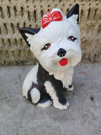 A white and black dog figurine is positioned centrally, seated on a textured grey concrete surface in the city of Sungurlare, Bulgaria. The figurine features a predominantly white head with black ears, prominent black eyes with red inner corners and stylized black eyelashes, a small black nose, and an open mouth revealing a bright red tongue. A red bow with white stripes adorns the top of its head between the ears. The body of the figurine is patterned with irregular black and white patches. In the background, a light brown or beige textured wall with a repeating pattern of small, vertical indentations extends across the frame. The lighting suggests an outdoor environment under natural daylight. No people or other active subjects are present.