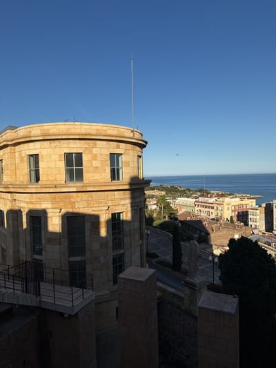 A sunlit, elevated view in Tarragona, Spain, showcases the city's architecture and coastal setting. A multi-story, cylindrical stone building dominates the left of the frame, with squared windows and shadowed areas. Adjacent to it, a stone wall with rectangular pillars overlooks the cityscape. Beyond the wall and building, a cluster of residential and commercial buildings with varied rooftops stretches towards the Mediterranean Sea. A clear blue sky with few visible clouds fills the upper portion of the image. A flagpole extends from the top of the stone building.