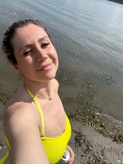 A woman in a yellow bikini is taking a selfie by the water. She is looking at the camera and smiling. The water is calm and there are some plants growing in it. The sand is visible in the foreground and there is a can of soda in her hand. The image appears to have been taken on a sunny day. It is not possible to determine the location as Pravda, Russia, from the image.