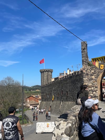 This image depicts Hostalric Castle in Hostalric, Spain. The castle features stone walls, a square tower, and a round tower with a red flag. Tourists are walking along the castle walls, and others are on the path below. The sky is blue with some clouds. A powerline runs through the sky, and a small airplane contrail is visible.