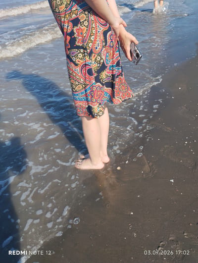 A person stands partially submerged in shallow ocean water on a sandy beach, holding a cell phone in their right hand. The individual is wearing a colorful, patterned dress. The scene is captured from a low angle, focusing on the lower body and the water's edge. The sand is dark and wet, with small shells scattered across it. Waves are gently lapping at the shore, creating foam and ripples. A shadow of the person is cast on the wet sand to the left. In the background, another person's legs are visible in the water, kicking up a splash. The image is dated "03.07.2026 17:02" and watermarked "REDMI NOTE 12". The location is the city of Abilyatag, Azerbaijan.
