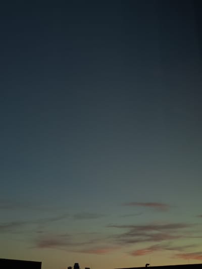 The image shows a twilight sky over a city. The sky is a gradient of deep blue at the top fading to lighter blues and hints of orange and pink near the horizon. Thin, wispy clouds are visible, tinged with pink from the setting sun. Silhouetted against the horizon are the rooftops of buildings, with some indistinct structures and antennas. The overall impression is a quiet urban evening.