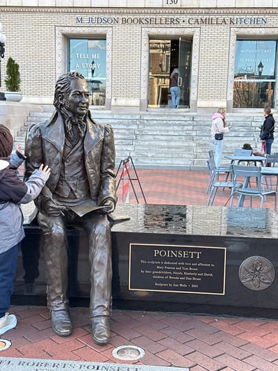 A bronze statue of Joel Roberts Poinsett sits on a bench in front of M. Judson Booksellers - Camilla Kitchen in Greenville, SC. A plaque notes it's dedicated to Mary Frances and Tom Bruce by their grandchildren; sculpted by Zan Wells in 2001. The bookstore's window displays "Tell Me a Story." Above, another window reads, "A party without cake is really just a meeting." People are visible on the steps behind the statue, and several gray metal chairs are placed nearby.