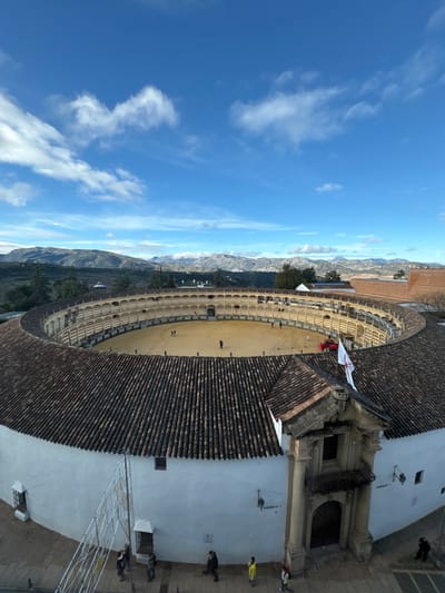 The image provides a high-angle view of the Plaza de Toros de Ronda, the bullring located in Ronda, Spain. The central feature is the bullring's circular sand arena, light tan in color, where approximately 10-15 individuals are dispersed, walking or standing. A small red utility vehicle is parked near the arena's right edge. Surrounding the arena are several tiers of seating, characterized by light-colored walls, archways, and supporting columns.

The bullring's structure includes a multi-pitched dark brown tiled roof that covers the spectator areas. The exterior wall visible to the viewer is white, presenting a prominent, ornate entrance (Puerta Grande) on the right side. This entrance features two large columns, a dark wooden double door, and a small balcony above it, topped with a tiled roof. A white flag with a red cross-like emblem is affixed to a pole above this entrance. Another entrance, labeled "PUERTA 1," is visible further to the left along the white exterior wall.

At street level, in front of the white wall, several individuals are observed walking on a paved surface. A decorative white light structure or archway is partially visible on the far left. The background reveals a clear blue sky with scattered white cumulus clouds. A vast range of rugged mountains, some appearing rocky and others vegetated, stretches across the distant horizon. Sparse green trees and other buildings with reddish-brown tiled roofs are visible between the bullring and the mountain range.