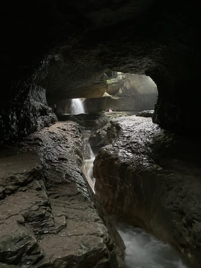 The image depicts the interior of a cave with a narrow passage carved by a stream. The walls of the cave are rough and wet, with visible layers of rock. Water flows rapidly through the passage, creating a frothy white appearance. In the background, a faint light illuminates the scene, suggesting an opening to the outside. There are two small figures visible in the distance, one standing on a ledge and another near a waterfall. The environment is damp and appears to be a natural geological formation. This scene is likely located in the vicinity of the Salta village in Russia, known for its numerous canyons and waterfalls.