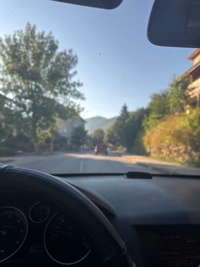 This is a first-person view from inside a vehicle, looking forward through the windshield. The car is driving on a road in a town, with buildings and trees lining the street. The sun is shining, casting a bright light on the scene. The road appears to be paved and is clear of obstructions. In the distance, a red car is visible, moving in the same direction. The dashboard of the car is partially visible in the foreground, with the steering wheel, speedometer, and a digital display showing information such as range and fuel consumption. The environment suggests a rural or suburban setting, likely in Smilyan, Bulgaria, given the information provided. The overall impression is of a sunny day drive through a quiet town.