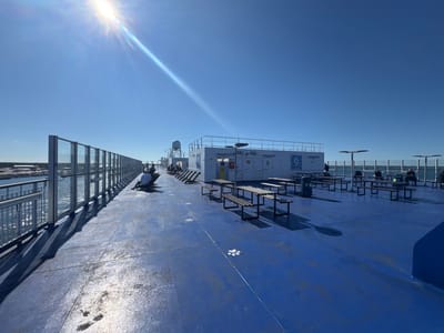 The image depicts the open deck of a ferry, likely docked or at sea near Karlskrona, Sweden, under a clear blue sky. Sunlight streams in from the upper left, casting long shadows across the bright blue deck. Several picnic tables with attached benches are scattered across the deck, some empty and others occupied by a few individuals.
To the left, a railing and a glass barrier separate the deck from the calm water, where a hint of land and structures can be seen in the distance, suggesting proximity to shore or a harbor. The ferry's white superstructure, featuring communication equipment like radar dishes, is visible in the background. White paw prints are painted on the deck at regular intervals.
On the deck, a few people are seated on benches or lounge chairs, some looking out at the water, others engaged in conversation or reading. The scene is orderly and serene, characteristic of a ferry crossing on a sunny day. The presence of the paw prints might indicate a designated area for pets or a playful design element.