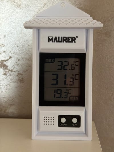 An indoor thermometer is displayed on a table. The thermometer is white and shaped like a small house with a peaked roof. The display shows three temperature readings: "MAX 32.6°C", "31.3°C", and "MIN 19.3°C". Below the display are two buttons labeled "CLEAR" and "°C/°F". The background is a textured wall.