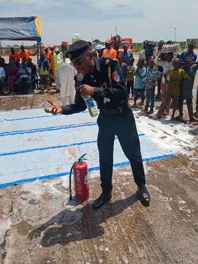 A man wearing a black uniform and a black beret with sunglasses is speaking into a microphone while holding a bottle of water in his right hand. He is standing on a concrete surface covered in white foam, next to a red fire extinguisher. In the background, a group of people are gathered, some are sitting and some are standing. There is a blue tarp to the left of the scene. The man is likely giving a speech or demonstration. This scene is located in Katabu, Nigeria, at the coordinates 10.71 latitude and 7.514 longitude.