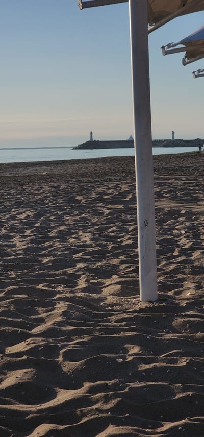 This media file depicts a beach scene with a focus on sand dunes. The sand is dark and textured with prominent ripples, suggesting it may have been recently exposed to wind or water. Small shells are scattered across the sand. A light-colored, horizontal metal bar cuts across the foreground, obscuring a portion of the sand.

In the background, a body of water, likely the sea, is visible. Beyond the water, a stone jetty or breakwater extends into the sea, with several lighthouses or similar structures situated along its length. The sky is clear and a pale blue, indicating a calm and sunny day. The lighting suggests it is either early morning or late afternoon, with the low angle of the sun casting long shadows across the sand.

The scene is likely located in Türkmenbaşy, Turkmenistan, as per the provided context. There are no visible people or specific activities taking place. The overall mood is tranquil and serene, characteristic of a beach at a quiet time of day. No text is visible in the image.