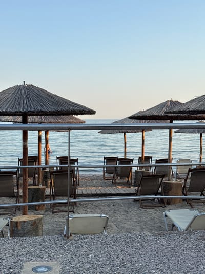 The image depicts a tranquil beach scene at Νέα Καλλικράτεια, Greece. The composition is dominated by rows of thatched umbrellas and empty lounge chairs, suggesting a prepared setting for beachgoers. The sea, with its gentle ripples and light blue hue, occupies the mid-ground, reflecting the clear, pale blue sky. A person can be seen in the water, partially submerged and appearing to be relaxing or floating. The foreground shows a pebbly ground, with some wooden planks forming a path. The overall atmosphere is serene and peaceful, likely captured during the late afternoon or early morning given the soft lighting and clear sky. There are no visible text elements in the image.