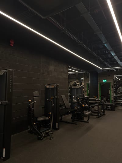 The image displays an unoccupied gym interior located in Dudley, United Kingdom. The environment exhibits a dark, industrial aesthetic, characterized by black block walls and a dark ceiling featuring exposed overhead utilities such as conduit, ductwork, and cable trays. The space is illuminated by multiple linear LED strip lights embedded in the ceiling. The floor is covered with dark, textured rubber matting.
On the far left wall, a red fire alarm call point is visible. The foreground and mid-ground are populated with various black-finished strength training machines, including a partial view of a "MATRIX" branded machine on the left, a seated chest press machine, and a leg curl/extension machine, all incorporating subtle orange accents on their adjustment mechanisms.
The right wall is partially covered by large, rectangular mirrors, reflecting sections of the gym and additional equipment, including weight racks and benches, further into the background. Towards the rear right, a dark door is visible, framed by a prominent bright green border and surmounted by an illuminated green "exit" sign with a left-pointing arrow. No individuals are present, and no actions or interactions are depicted.