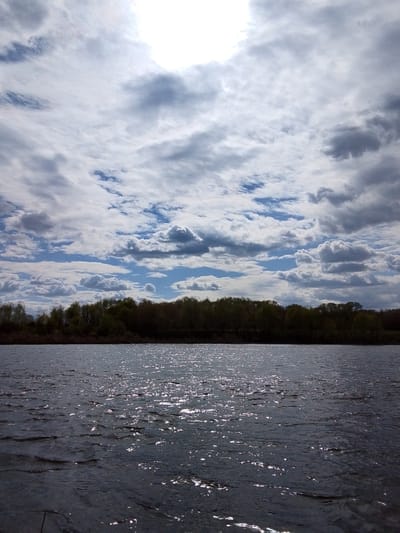 The image depicts a body of water, possibly a river or lake, under a cloudy sky. A line of trees is visible on the horizon across the water. Sunlight glints off the surface of the water, creating a sparkling effect. The location is Дерезовка, Russia.