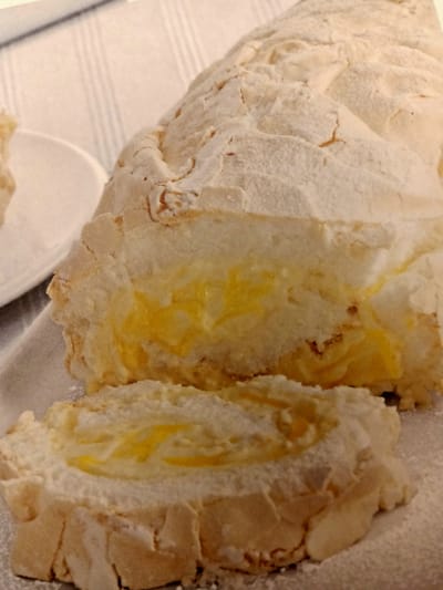A close-up, overhead shot shows a baked meringue roll filled with a bright yellow cream. The meringue exterior is light brown and powdered with white sugar. The roll is sliced, revealing the creamy filling. The roll sits on a white platter next to a white plate and a light-colored striped tablecloth. The scene is likely indoors, possibly in a home kitchen in Rumaithiya, Kuwait, given the context.